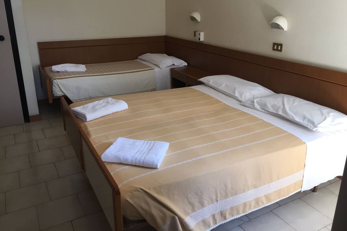 Habitación de hotel con dos camas individuales y una cama doble, todas con ropa blanca y cubrecamas a rayas. Se observan luminarias fijadas a la pared y un enchufe visible.