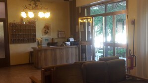 A reception area with a wooden counter, a chandelier, and a view of the outdoors through large windows. - Hotel Belvedere (Castrocaro Terme)