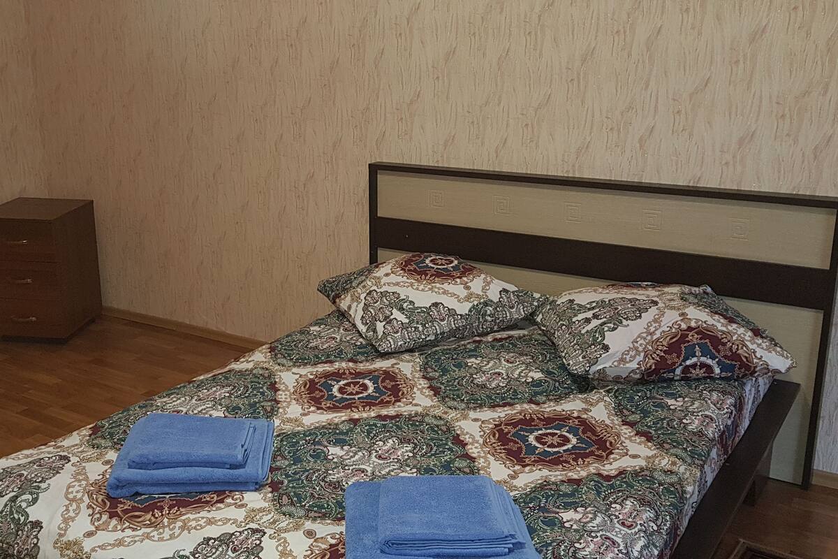 Un lit bien fait, avec une literie à motifs et deux serviettes pliées, une commode en bois et un plancher en bois, dans une chambre d’hôtel.