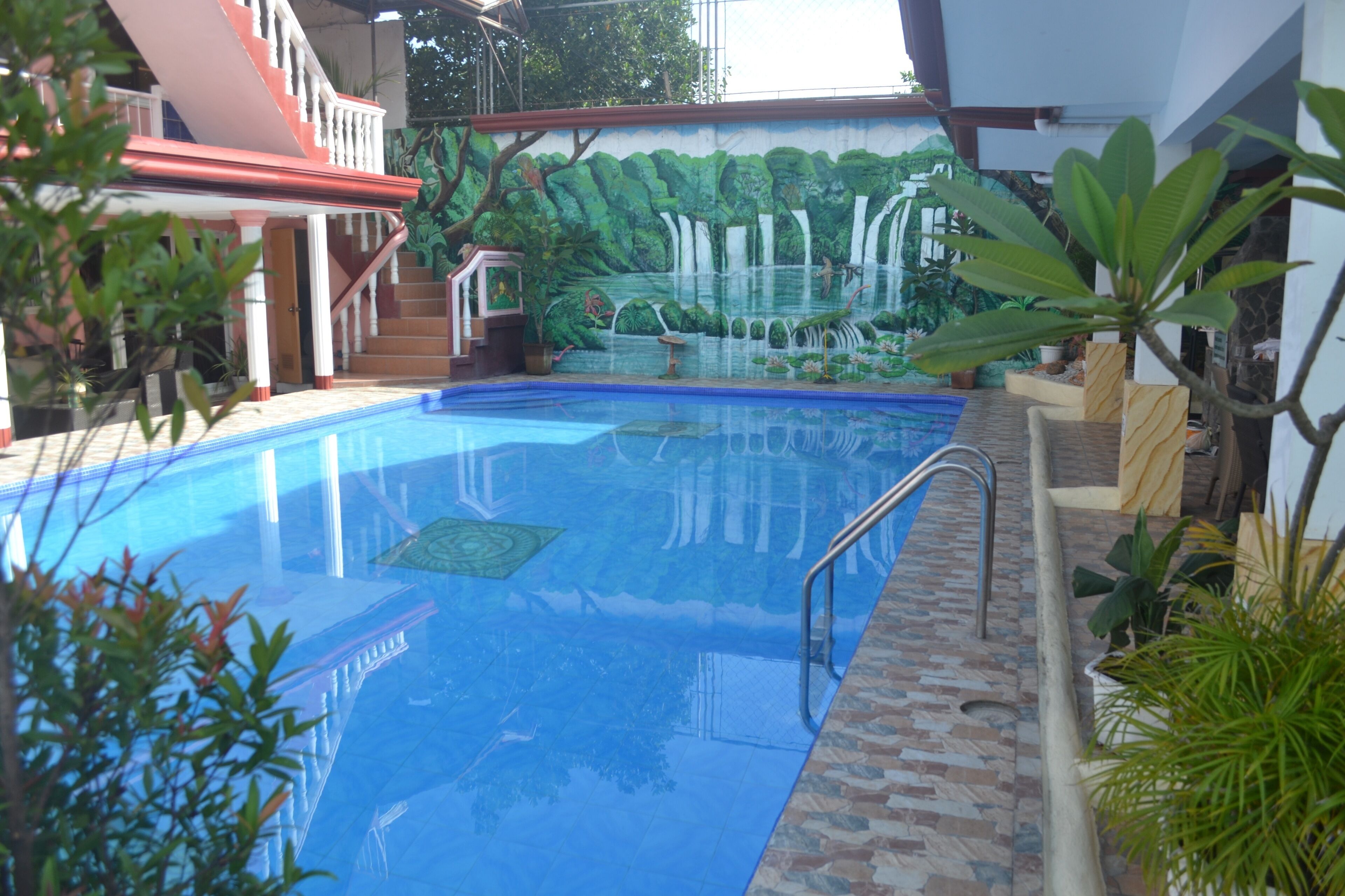 A swimming pool with a mural of a waterfall and greenery, surrounded by plants and a staircase.