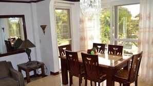 A dining area with a table set for six, surrounded by wooden chairs. There is a chandelier above the table, and the room has large windows with curtains.