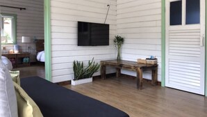 Ein Zimmer mit weißen Holzwänden, Holzboden, einer schwarzen Couch, einem Holztisch, einem an der Wand befestigten Fernseher und einer Tür mit grünem Rahmen.