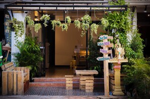 Fachada con plantas colgantes, bancos de madera y un cartel con indicaciones.