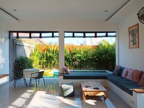 A modern living room with a glass coffee table, a sofa with cushions, a small wooden table, and a view of a swimming pool and greenery outside. - Fabia Villas (Seminyak)