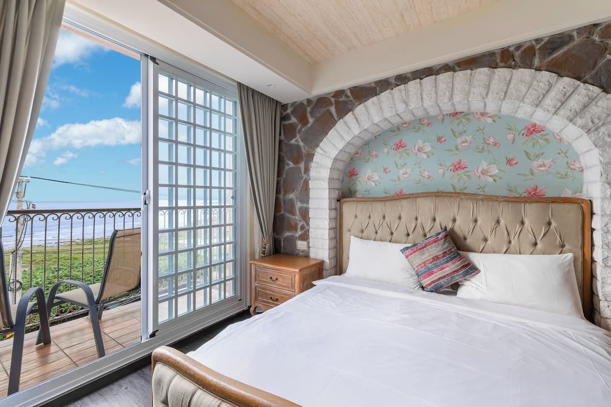 A bedroom with a large bed, a stone fireplace, a balcony with a view, and a wooden nightstand.
