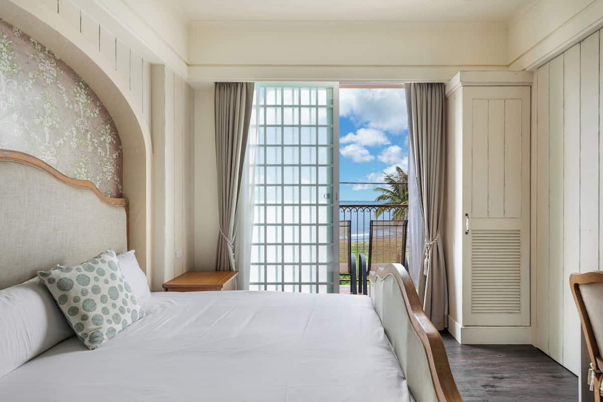 A hotel room with a large bed, a nightstand, a view of the outdoors, and a door leading to another room.