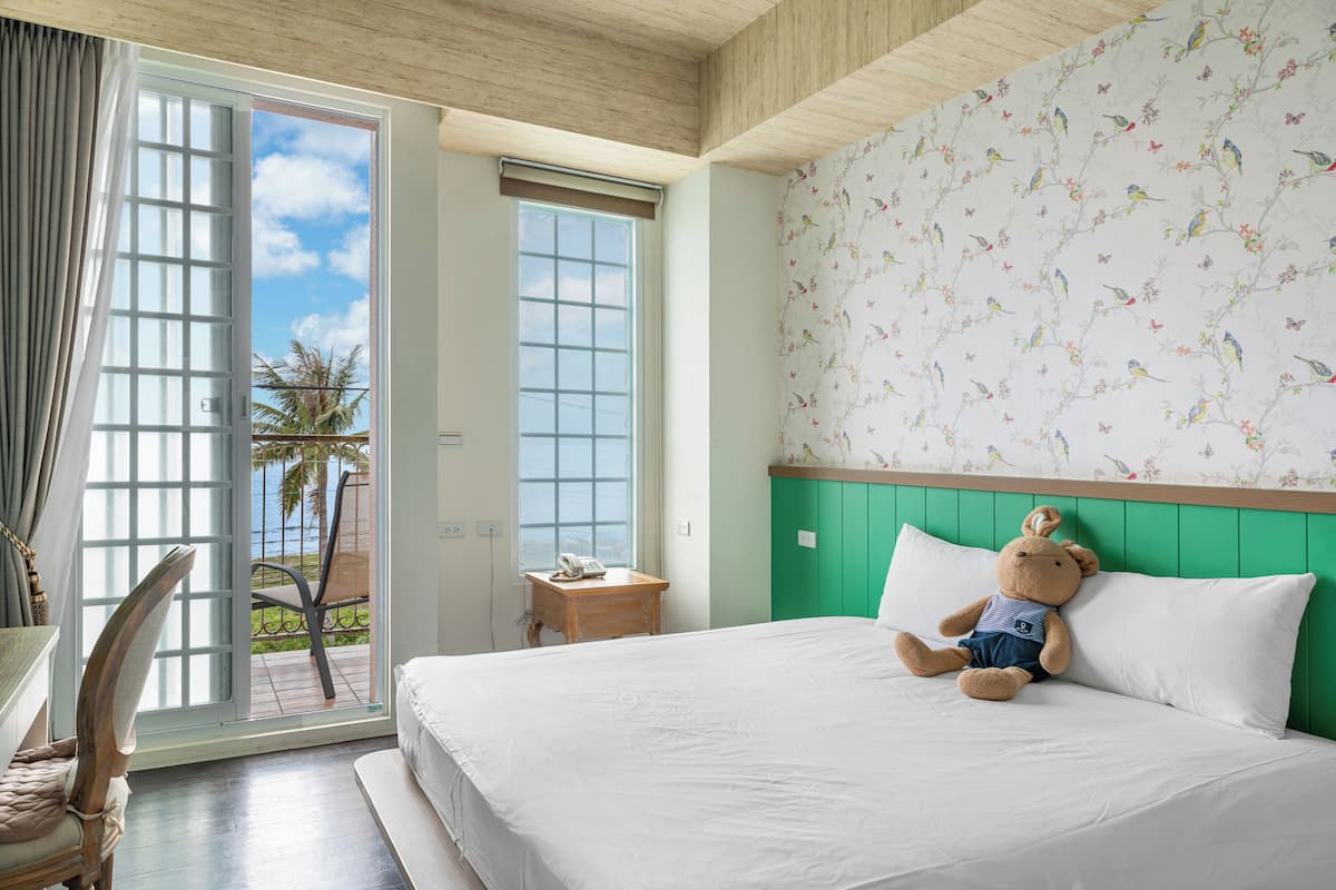 A hotel room with a bed, a chair, a small table with a phone, and a window with a view of palm trees.