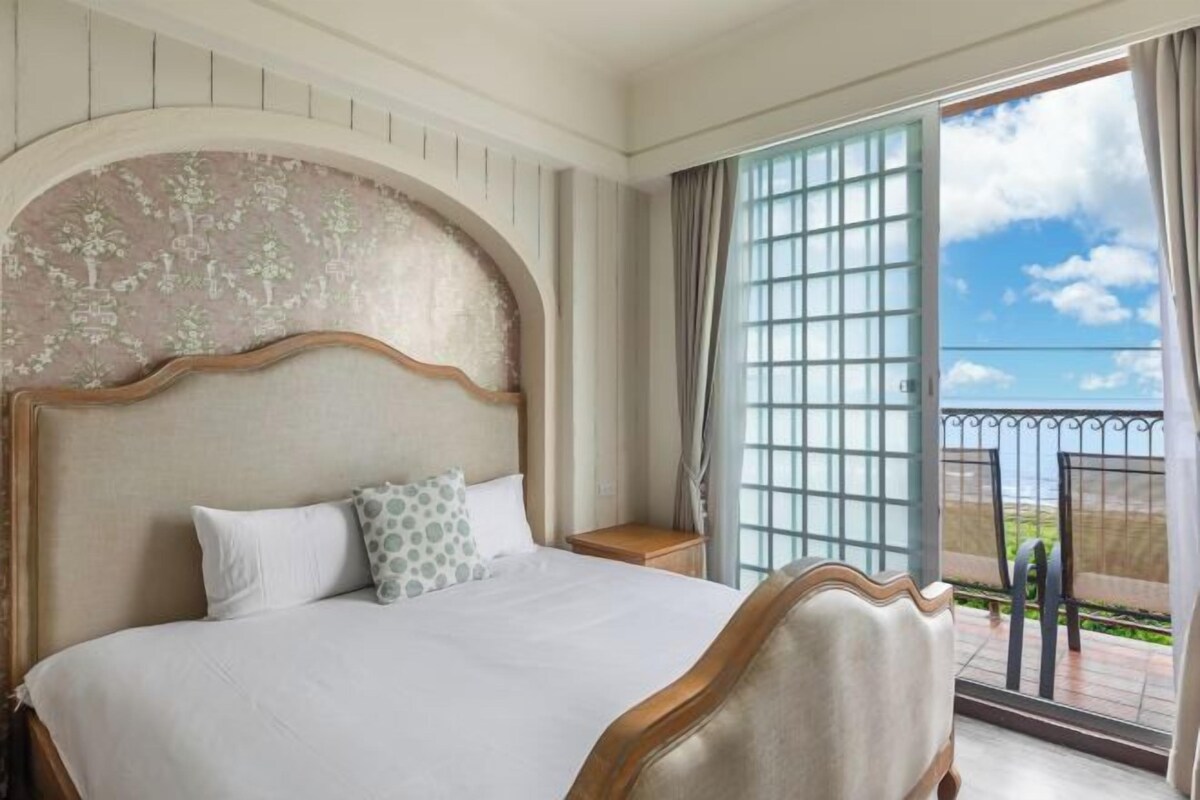 Een hotelkamer met een groot bed, een houten hoofdeinde, een nachtkastje en uitzicht op zee vanaf een balkon met een metalen hekwerk.