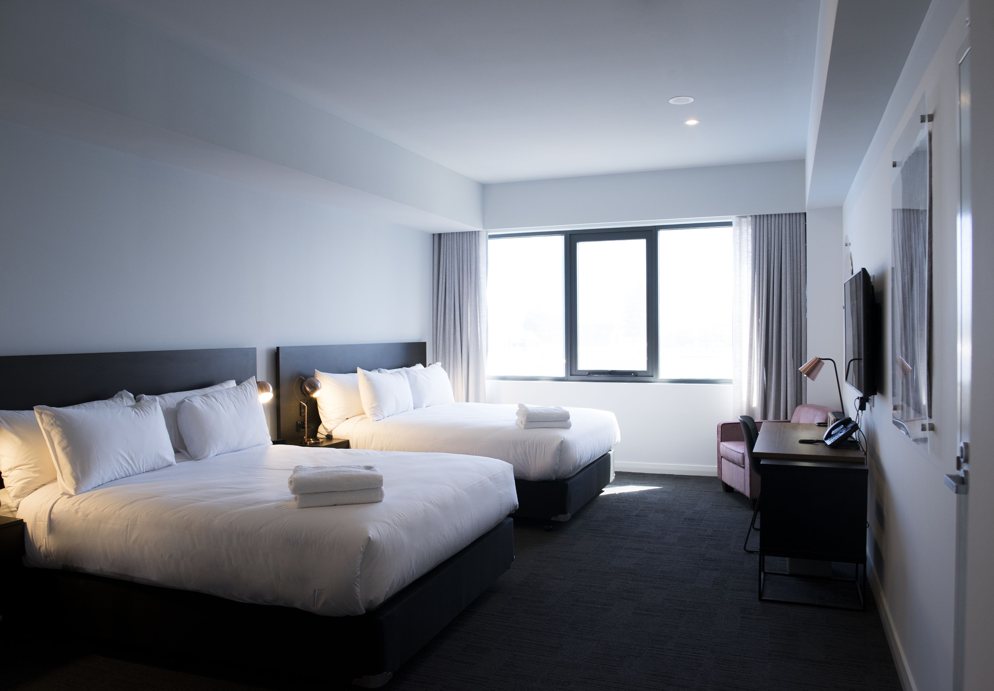 A modern hotel room with two beds, a desk, a chair, and a large window.