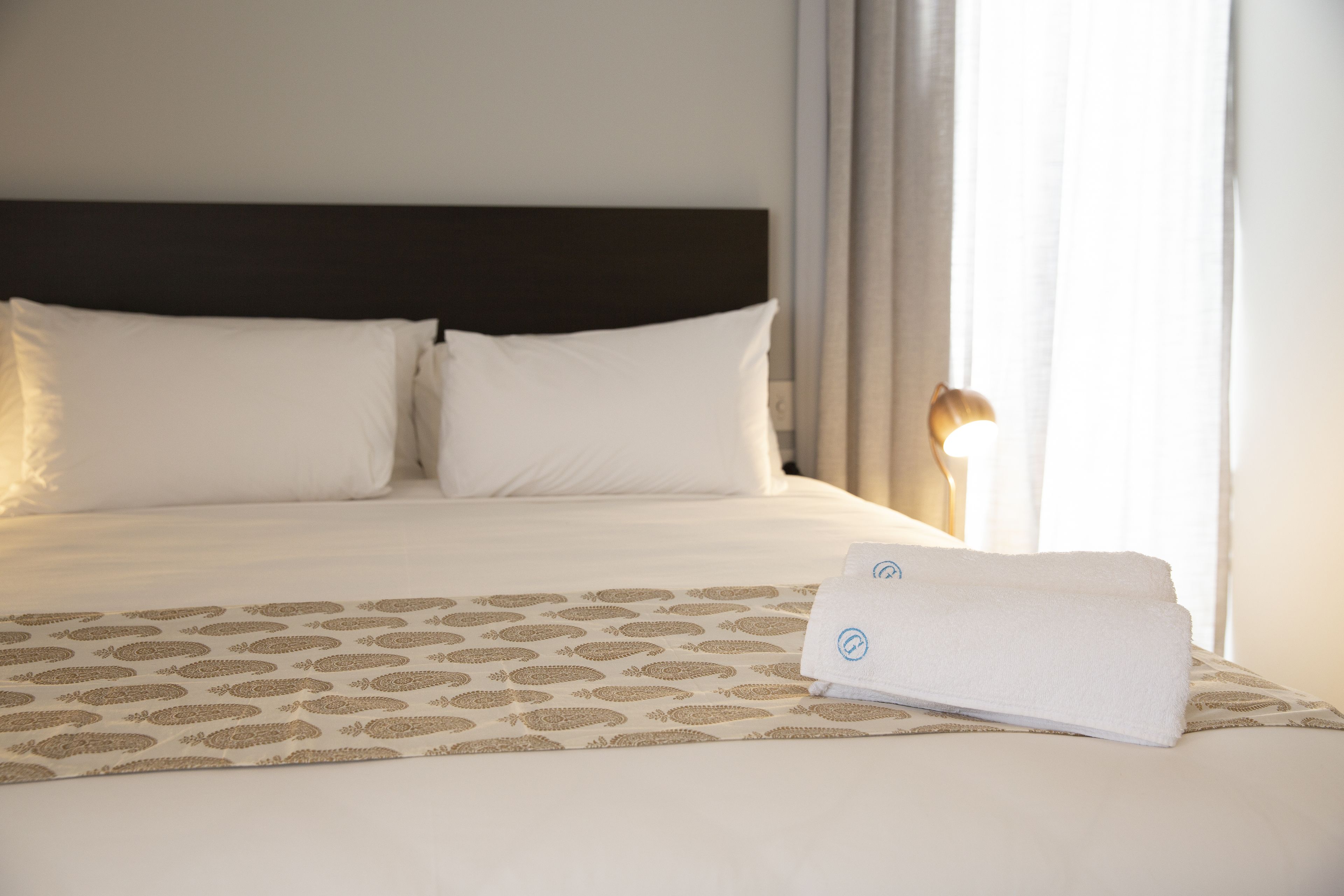 Een opgemaakt bed met witte lakens en een geborduurd sprei, een donker hoofdbord en een raam met gordijnen.