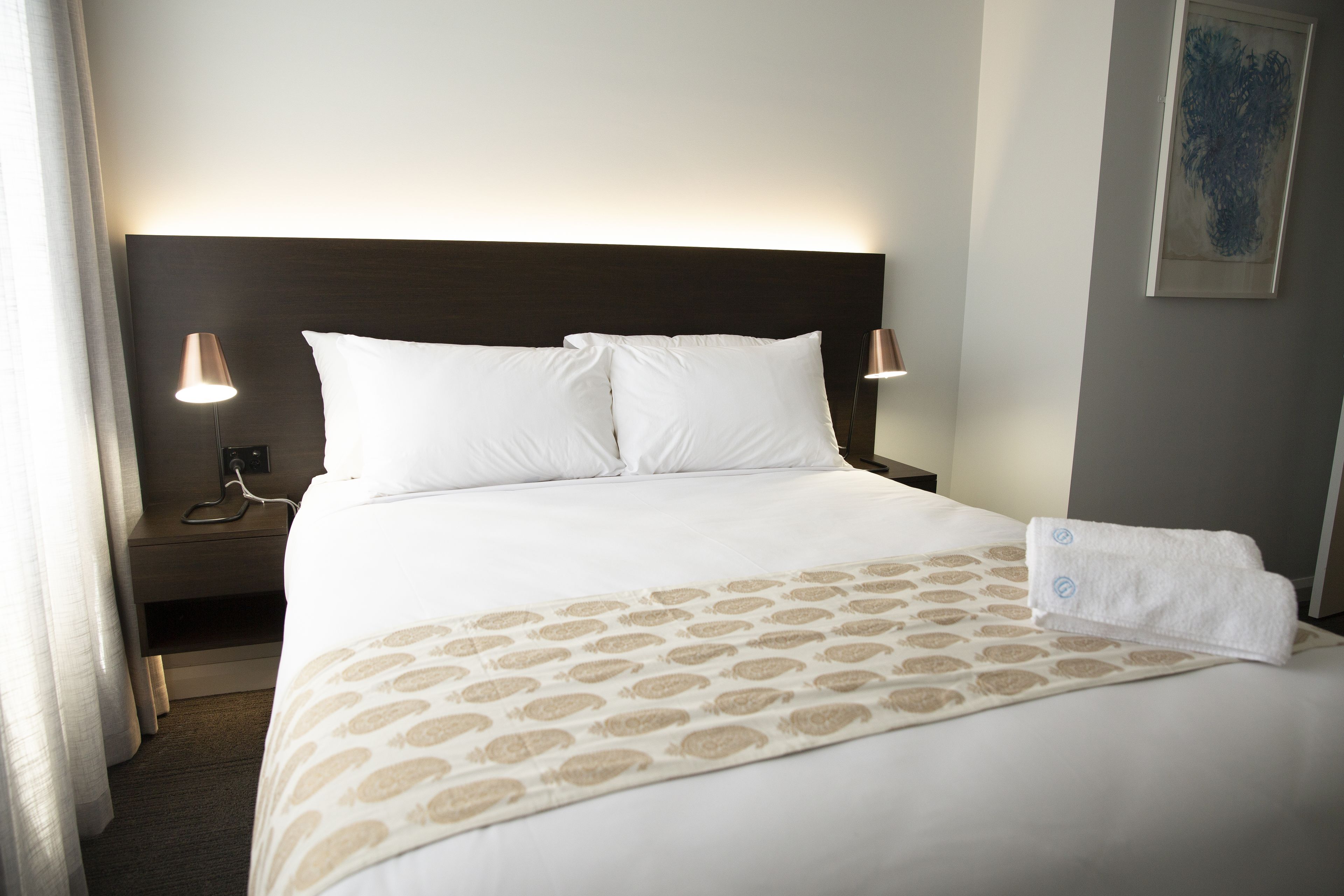 Una habitación de hotel con una cama bien tendida, dos lámparas de noche, una obra de arte en la pared y un enchufe visible en la mesita de noche.