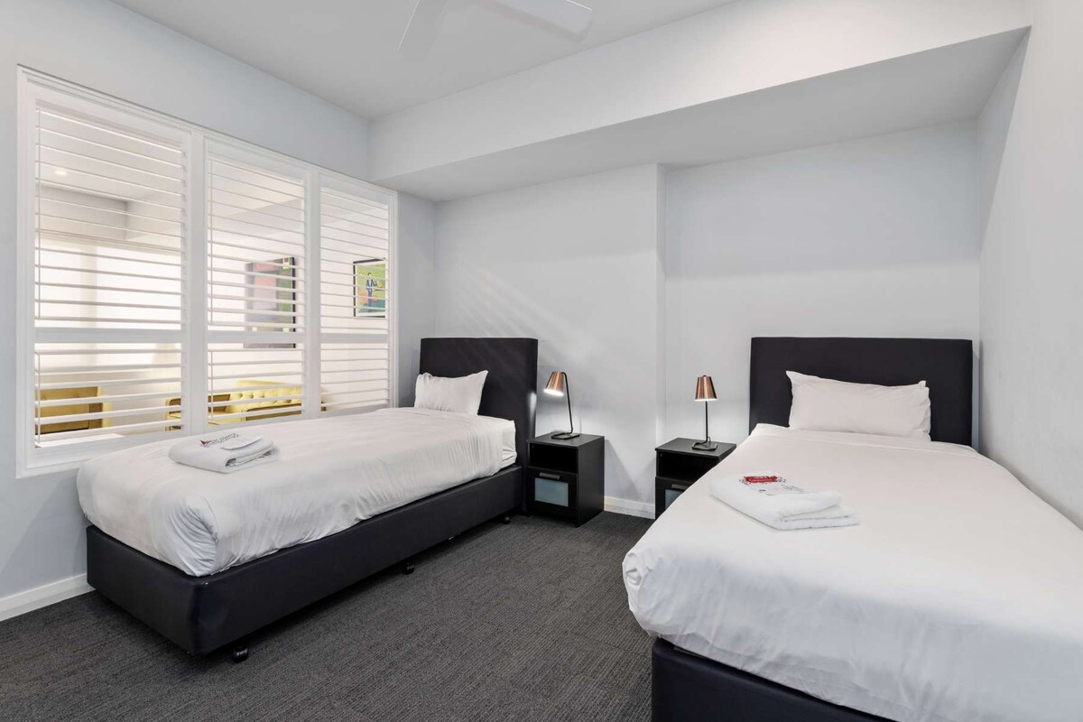 Habitación de hotel con dos camas, un ventanal con persianas, una mesita de noche con lámpara y piso alfombrado.