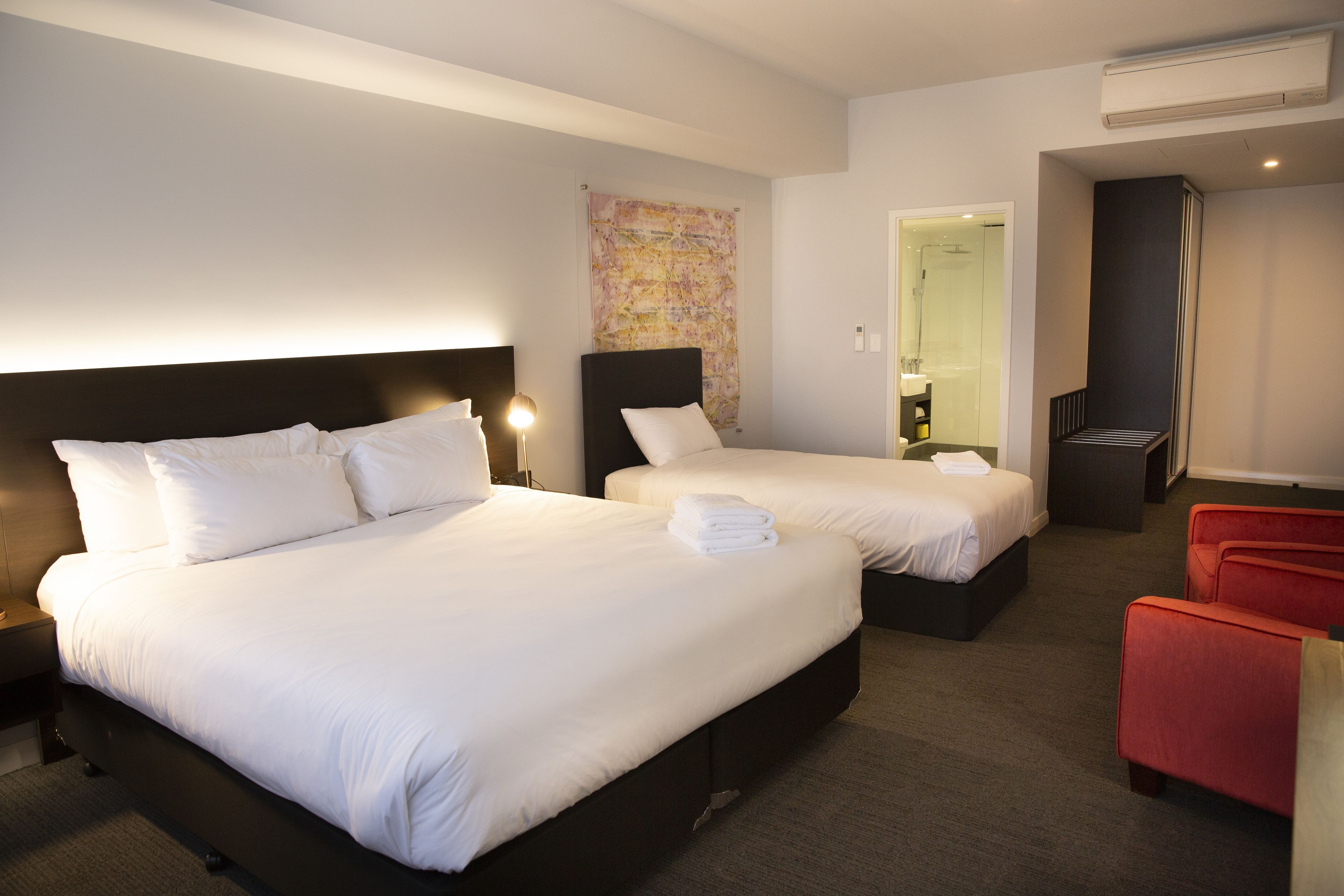 A hotel room with two beds, a red chair, and a bathroom visible through an open door.