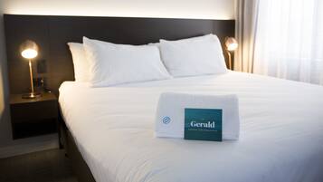 A neatly made bed with a folded towel and a hotel name tag, a nightstand with a lamp, and a window with curtains.