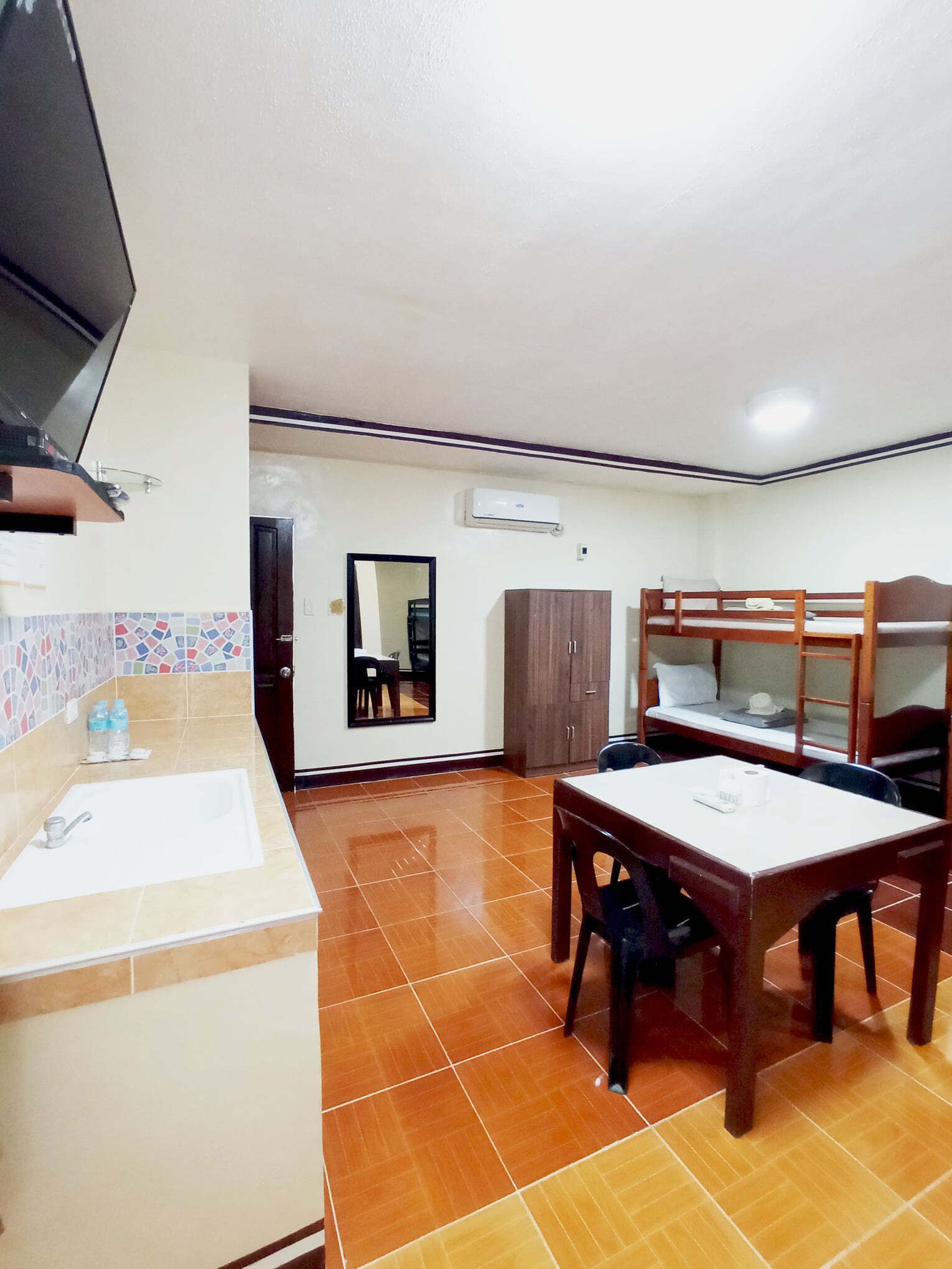 Una habitación con suelo de baldosas, lavabo, espejo, armario, literas y televisión.