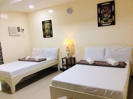 Quarto de hotel com duas camas de solteiro, uma mesinha de cabeceira com abajur, um relógio de parede e uma pintura floral emoldurada.