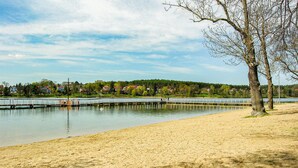 A sandy beach with a bridge over calm water, surrounded by trees and houses in the background.