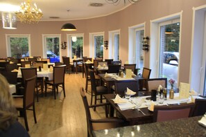 A restaurant with multiple tables and chairs, large windows, and a chandelier. - Hotel Haus Hubertus (Schneverdingen)