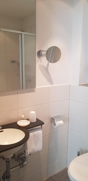 A bathroom with a sink, mirror, towel rack, toilet paper dispenser, and a shower area. - Hotel Haus Hubertus (Schneverdingen)