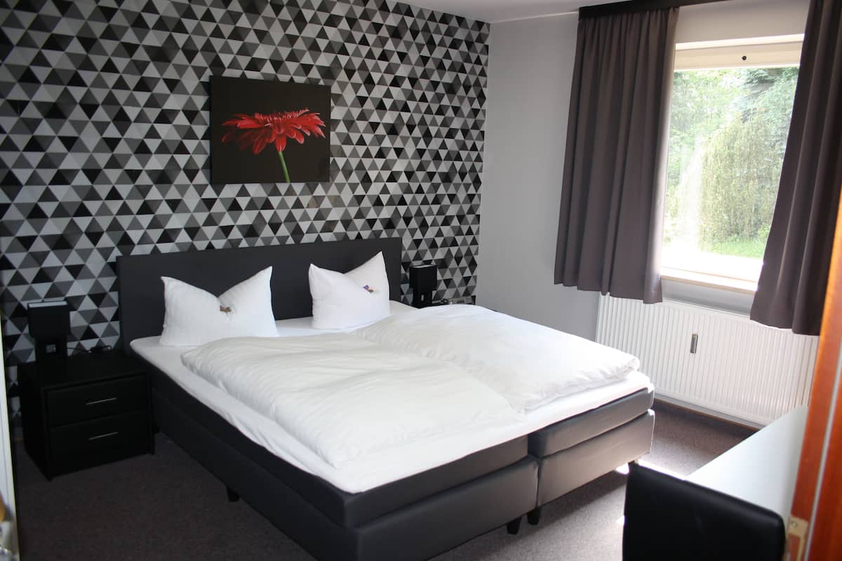 A modern bedroom with a geometric patterned wall, a large bed with white bedding, a nightstand, a bedside table, a black chair, and a window with curtains.