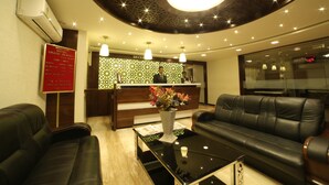 A hotel reception area with a reception desk, a black leather sofa, a coffee table, and a decorative vase. - Hotel Grandpragati (Surat)