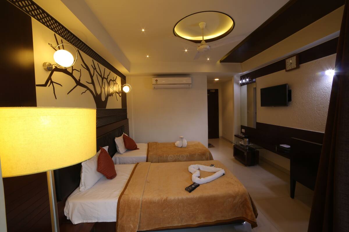 A hotel room with two beds, a bedside table, a lamp, a wall-mounted TV, and an air conditioning unit.