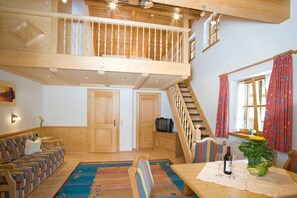 A spacious room with a high ceiling, a wooden staircase, a sofa set, a dining table with wine glasses, and a television.