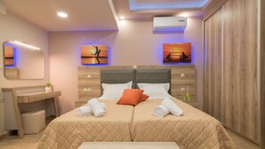 A double bed with a quilted beige cover, two pillows, and a red accent pillow. There is a wooden headboard, a bedside table with a plant, and a bench at the foot of the bed. - Zante Sky Suites (Zakynthos)