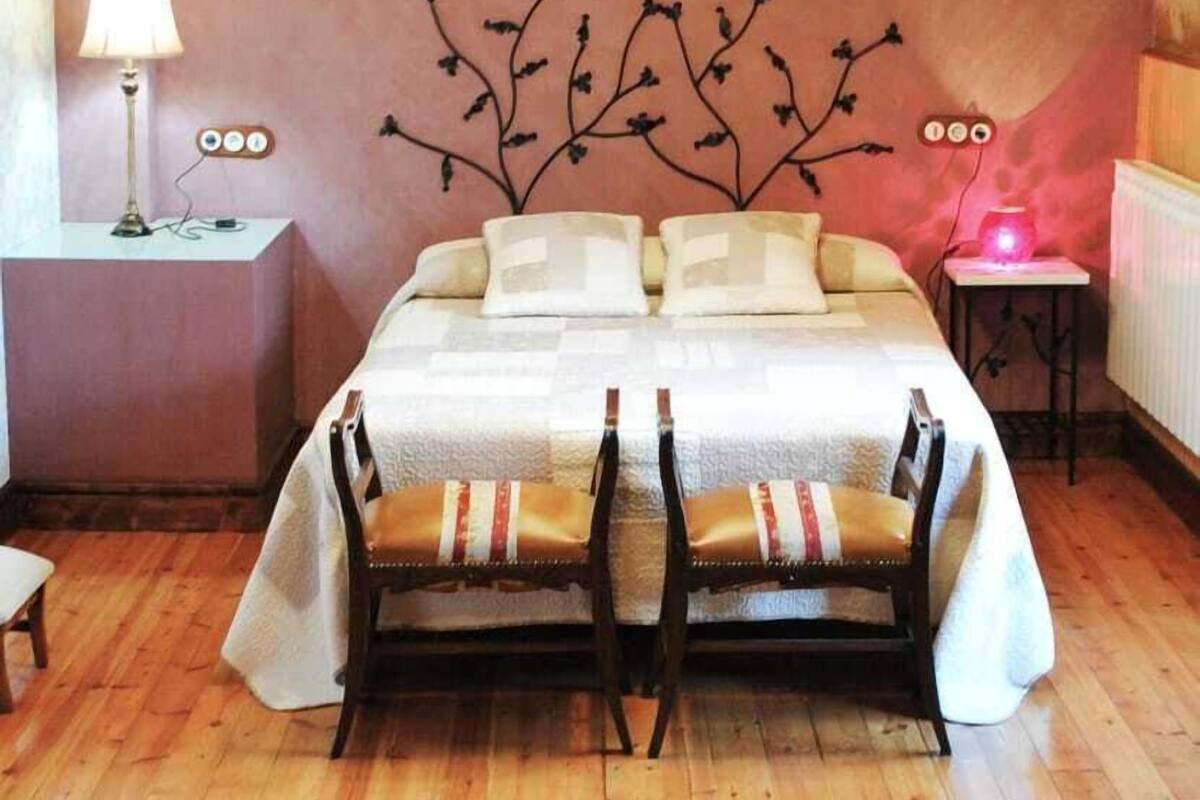 Un dormitorio con techo de madera, una cama con colcha blanca, dos sillas, una mesita de noche con lámpara y un adorno en la pared.