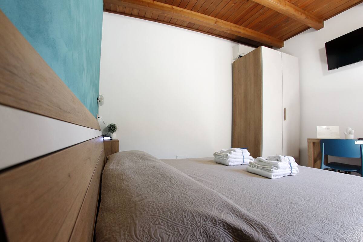 A bedroom with a wooden headboard, a bed with a patterned bedspread, a desk with a chair, and a television mounted on the wall.