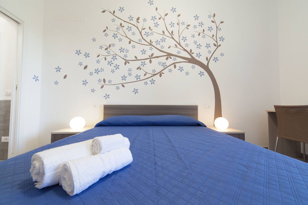 A neatly made bed with blue bedding and two rolled white towels, a wall decal of a tree with flowers, and two bedside lamps.