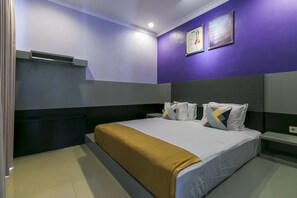 A modern hotel room with a large bed, a headboard, a bedside table, and a wall-mounted shelf.