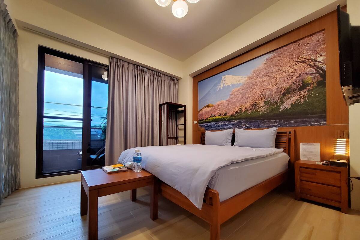A hotel room with a bed, a bedside table, a nightstand, a large window with curtains, and a painting of cherry blossoms.
