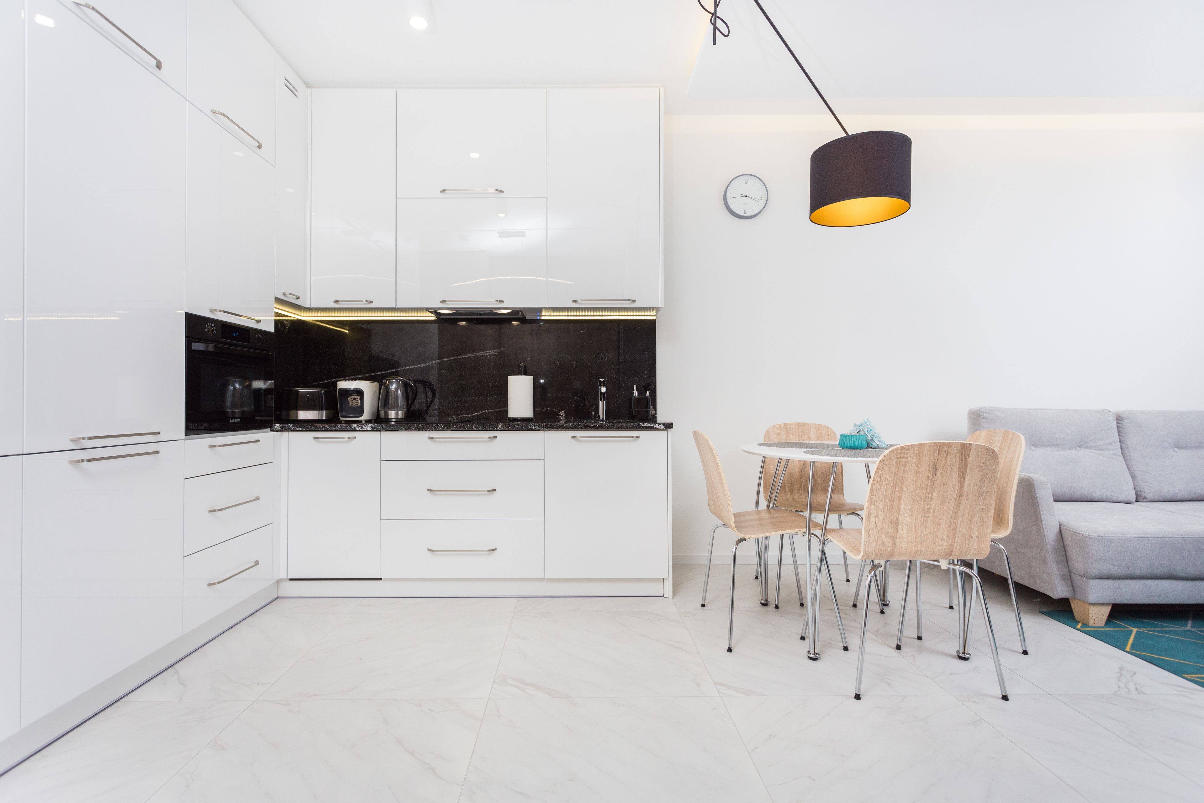 Une cuisine moderne avec des meubles blancs, un plan de travail noir et un coin repas comprenant un canapé gris et des chaises en bois.