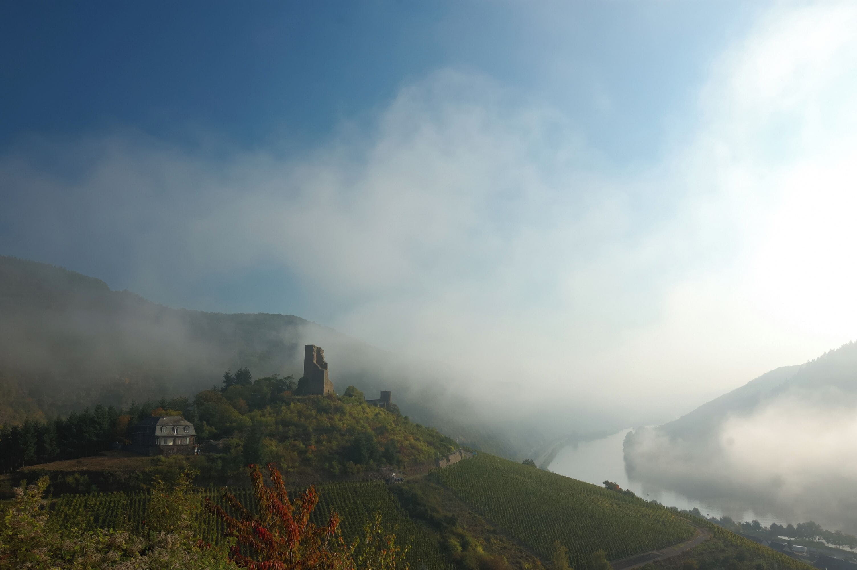 A misty landscape with a historic building on a hill, surrounded by lush greenery and a river.