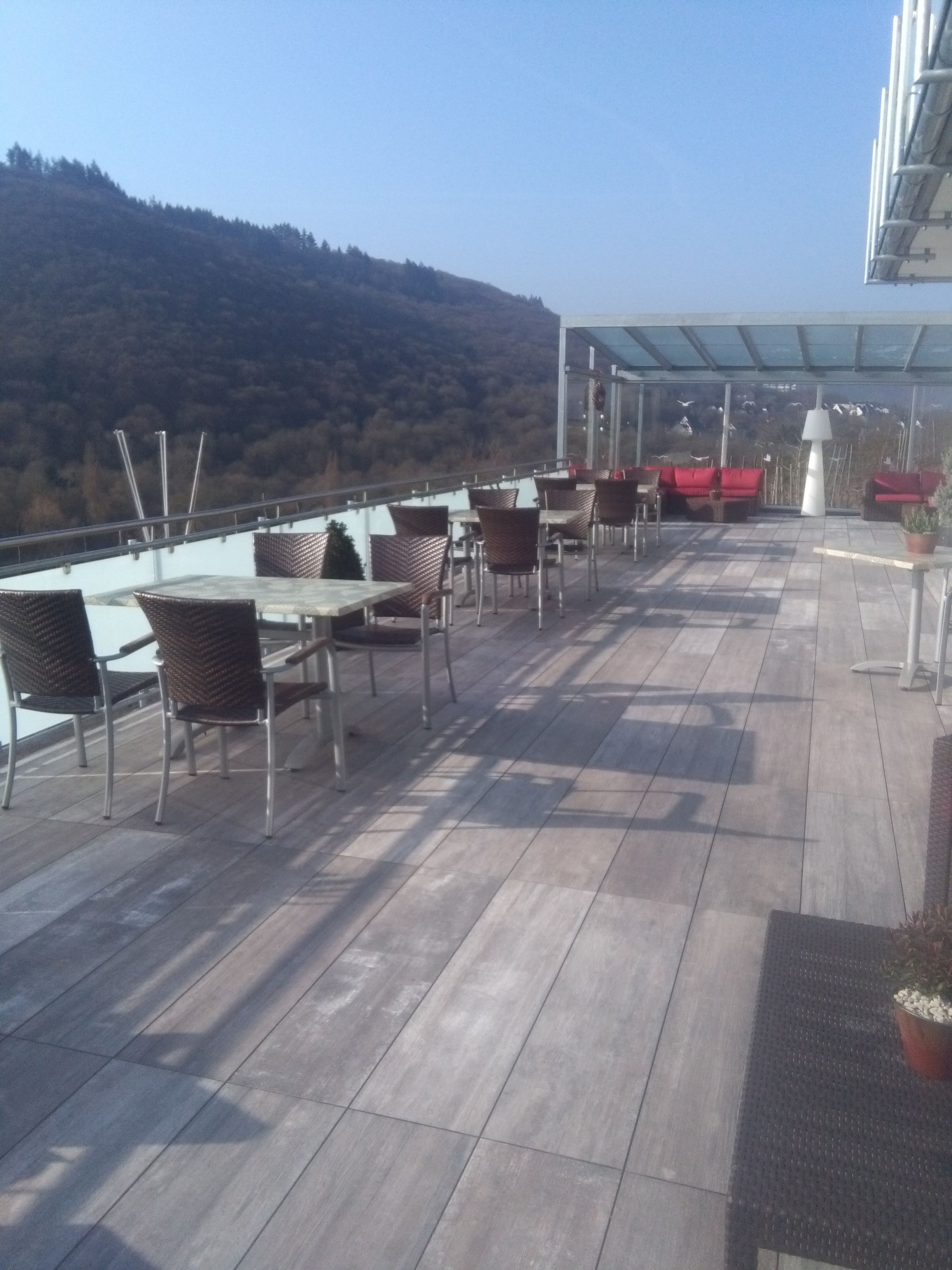 Una terraza al aire libre con mesas y sillas, un dosel de vidrio y vistas a una ladera boscosa.