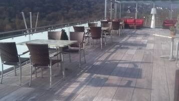 Una terraza al aire libre con mesas y sillas, un dosel de vidrio y vistas a una ladera boscosa.