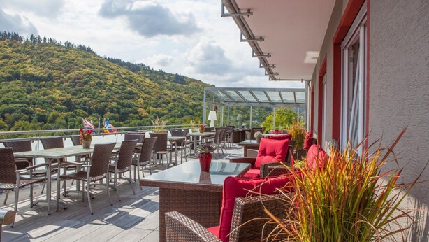 Een terras buiten met rieten meubels, een glazen tafel en rode kussens.