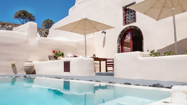 Un espace piscine avec une piscine à l’eau d’un bleu limpide, des murs blancs et une porte rouge. On y trouve des parasols et une table avec des chaises.