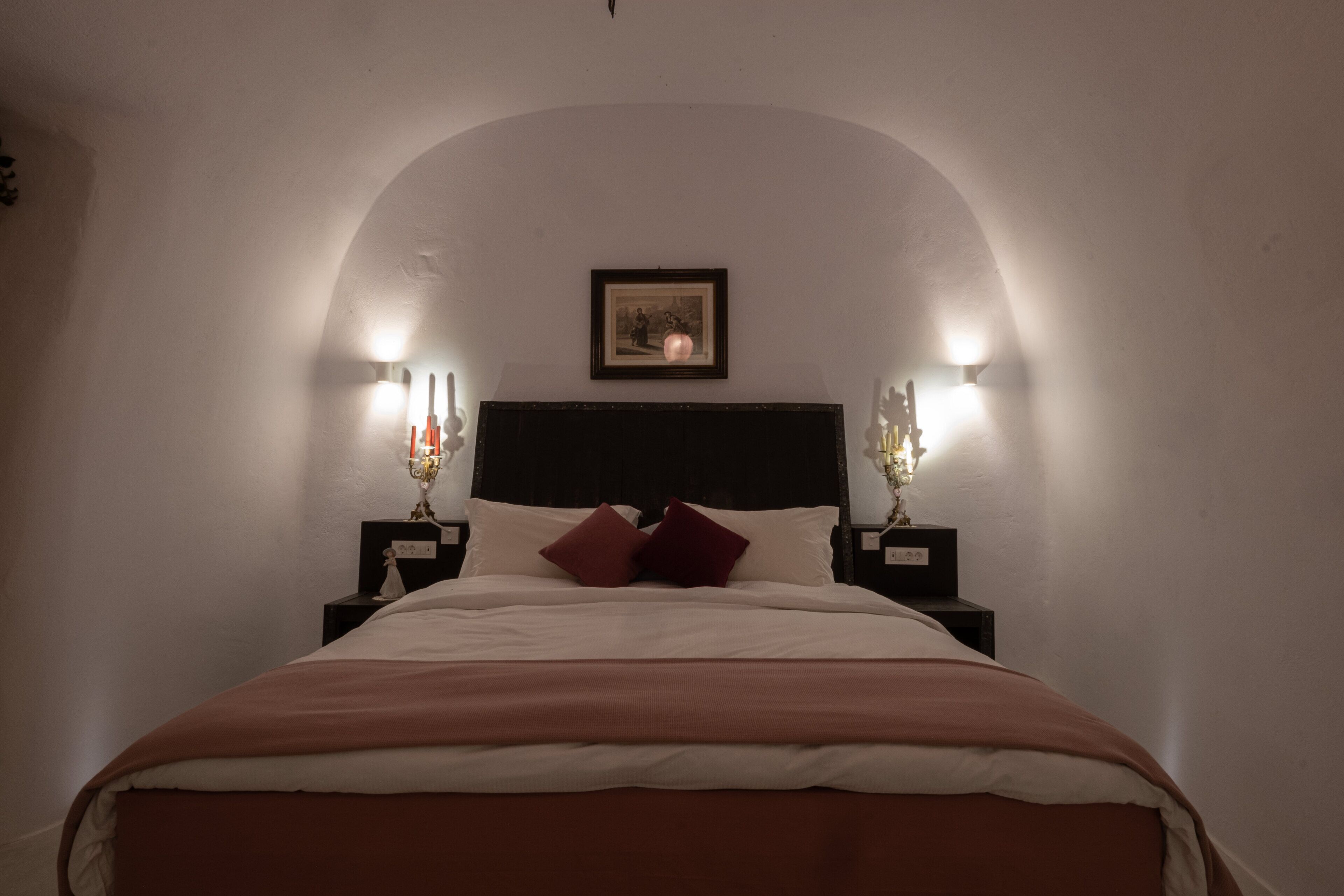 A bedroom with a large bed, two bedside tables with candles, a framed picture on the wall, and a white ceiling.