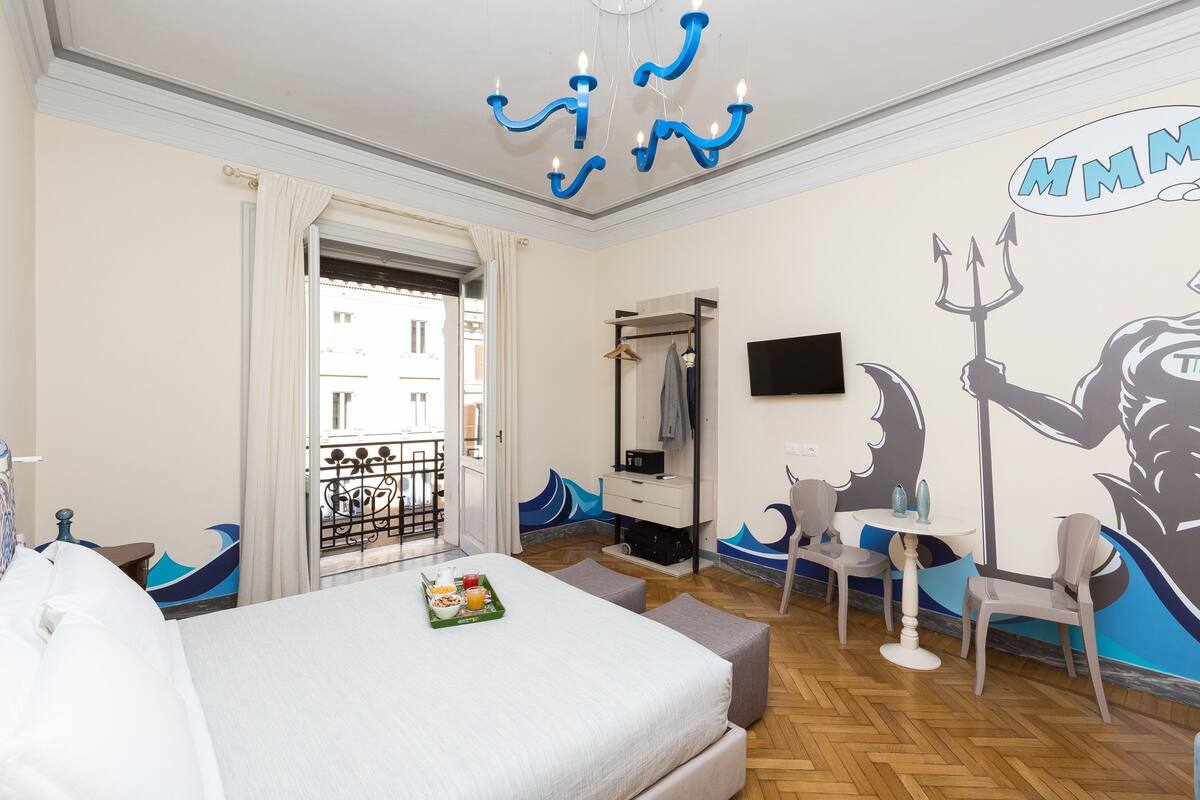 A hotel room with a bed, a dining table with chairs, a TV, and a mural on the wall.