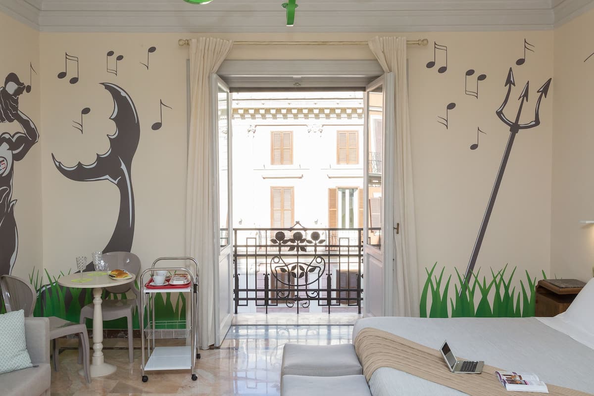 A hotel room with a bed, a desk with a laptop, a dining table with a tray, and a balcony with a view of buildings.