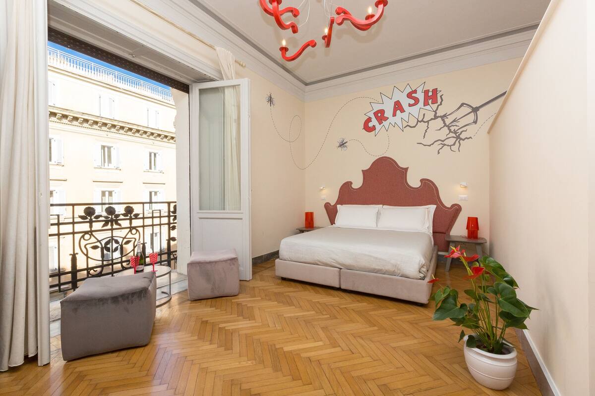 A bedroom with a bed, a potted plant, and a view of a building through a window.