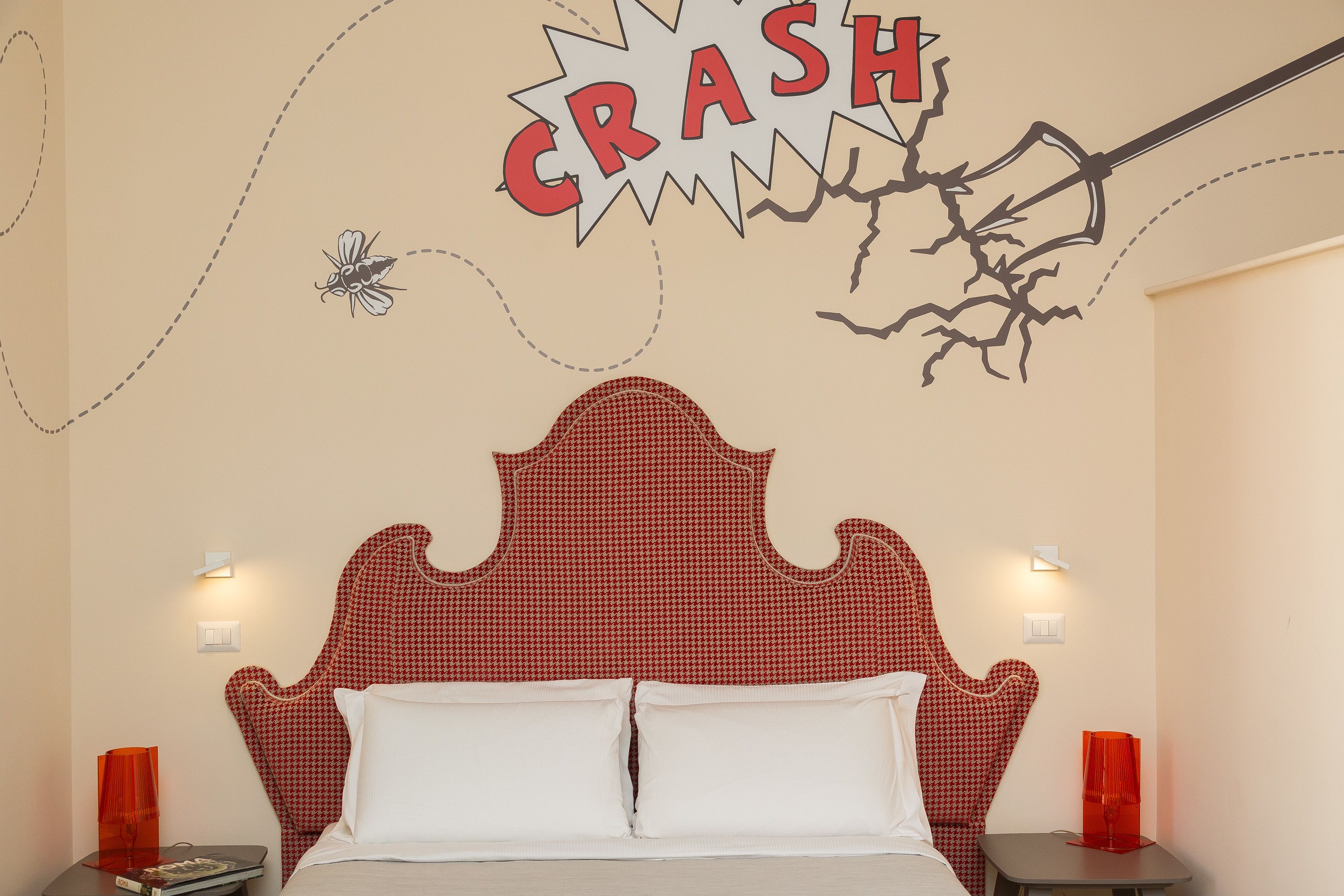 A bedroom with a red and white checkered headboard, two bedside tables with red lamps, and a wall decoration featuring a 'CRASH' comic-style explosion.