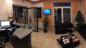 Een receptie met een balie, computer en een televisie. Er zijn twee zwarte banken, een salontafel met tijdschriften, en een kerstboom.