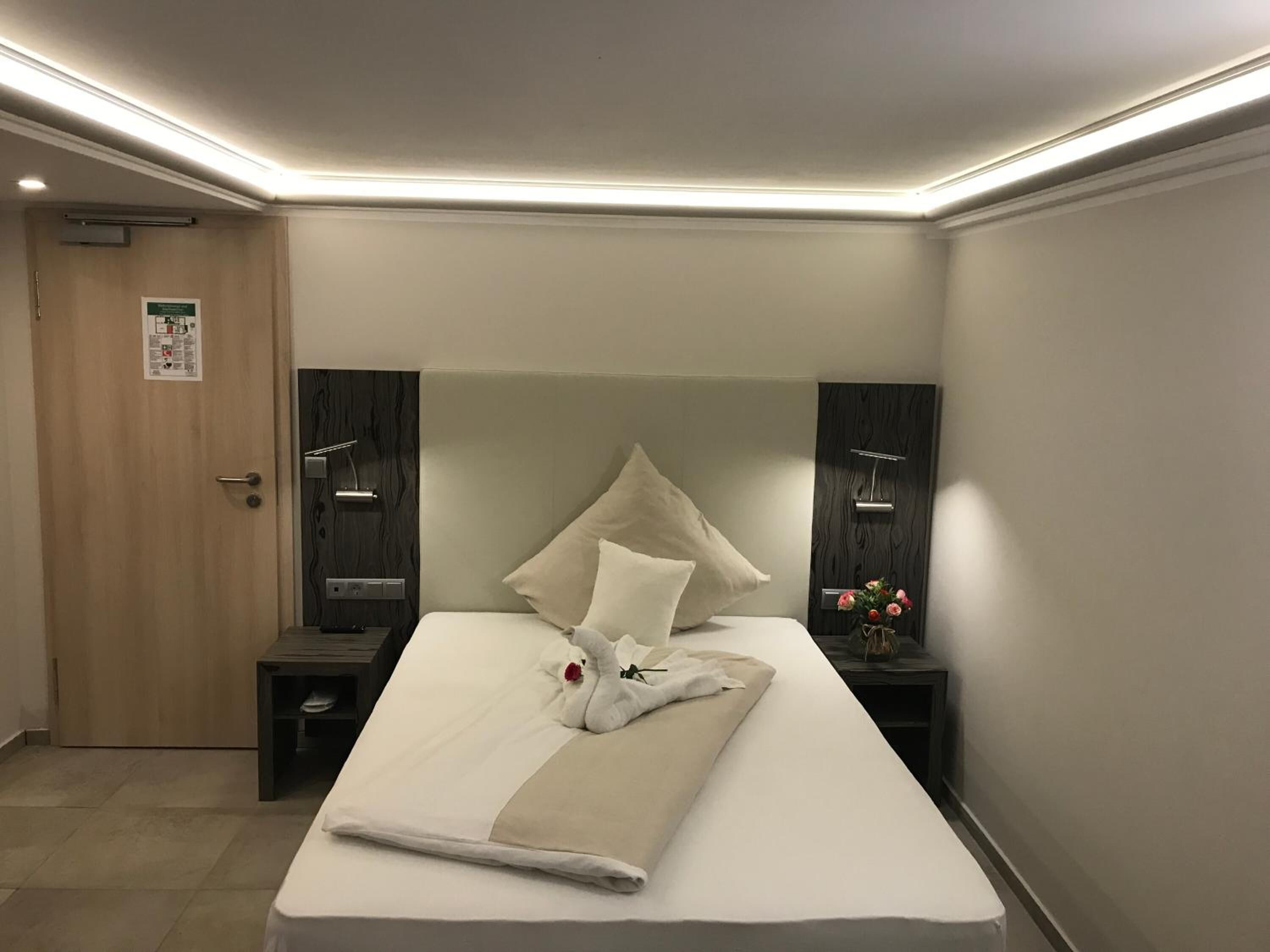 A modern hotel room with a neatly made bed, a wooden door, and a small bedside table with a lamp.