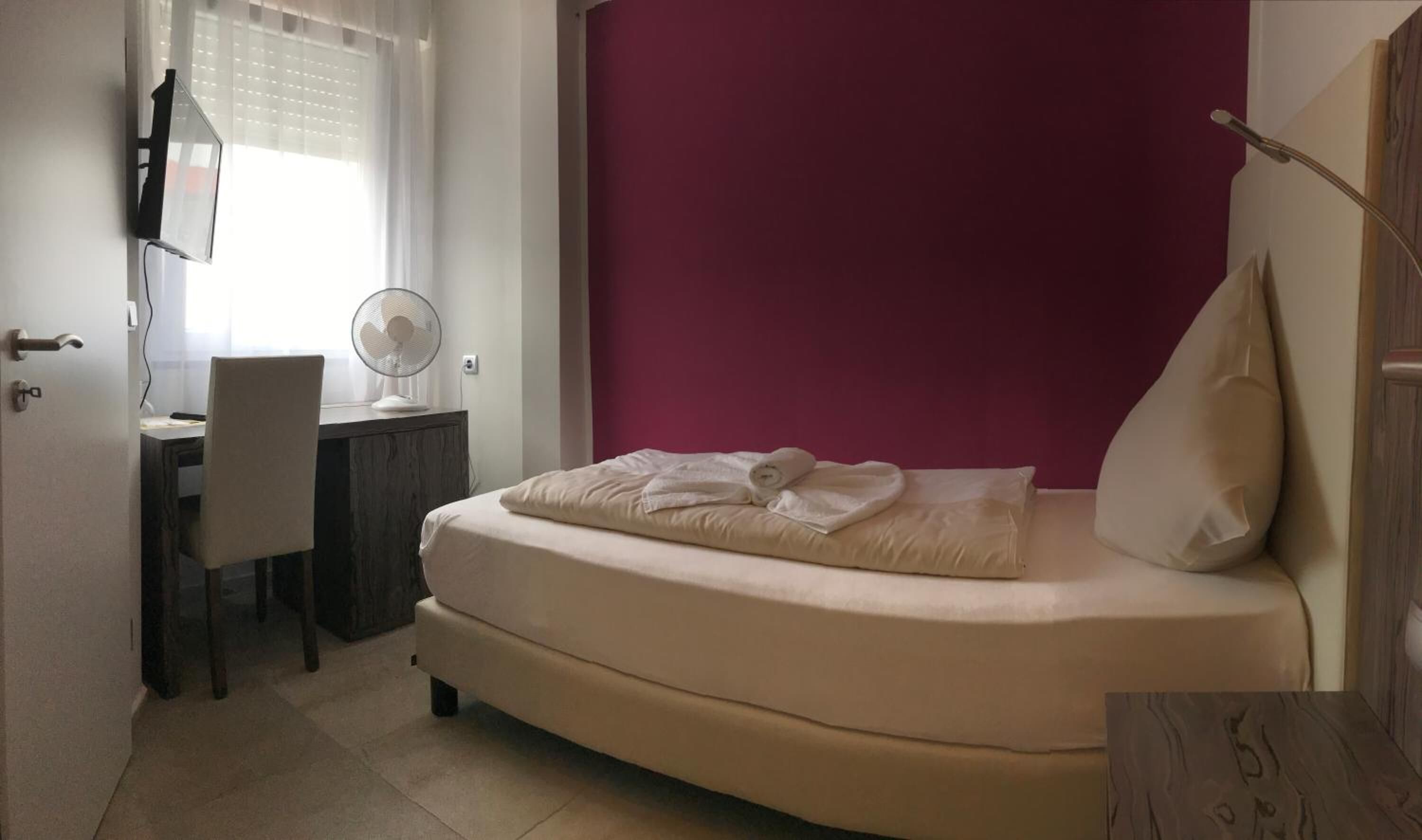 A bedroom with a bed, a desk, a chair, a fan, and a window with blinds.
