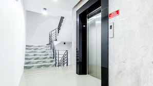 A modern elevator with a red safety sign, a marble-tiled wall, and a staircase with metal railings. - Napas Residence (Hat Yai)