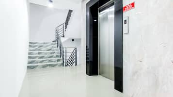 A modern elevator with a red safety sign, a marble-tiled wall, and a staircase with metal railings.