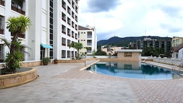 A swimming pool area with a tiled deck, surrounded by buildings with balconies and a mountain in the background.